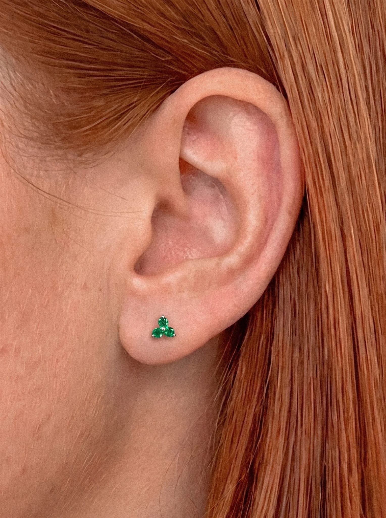 Nahaufnahme des Ohrs einer Person mit glattem rotem Haar, die den Ohrring „Trinity cz“ von Titanio-Noble Art trägt – ein kleiner grüner, dreieckiger Ohrstecker, der ein elegantes Piercing für alle darstellt, die einen raffinierten Stil schätzen.