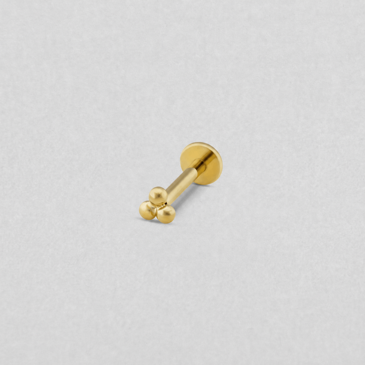 Découvrez le modèle « Trinity Liso » de Titanio-Noble Art : une petite boucle d'oreille de type labret en doré, ornée de trois perles rondes et lisses à l'avant et dotée d'un design moderne et discret, équipée d'un système de visage interne pour un port sûr et confortable, présentée sur un fond gris clair.