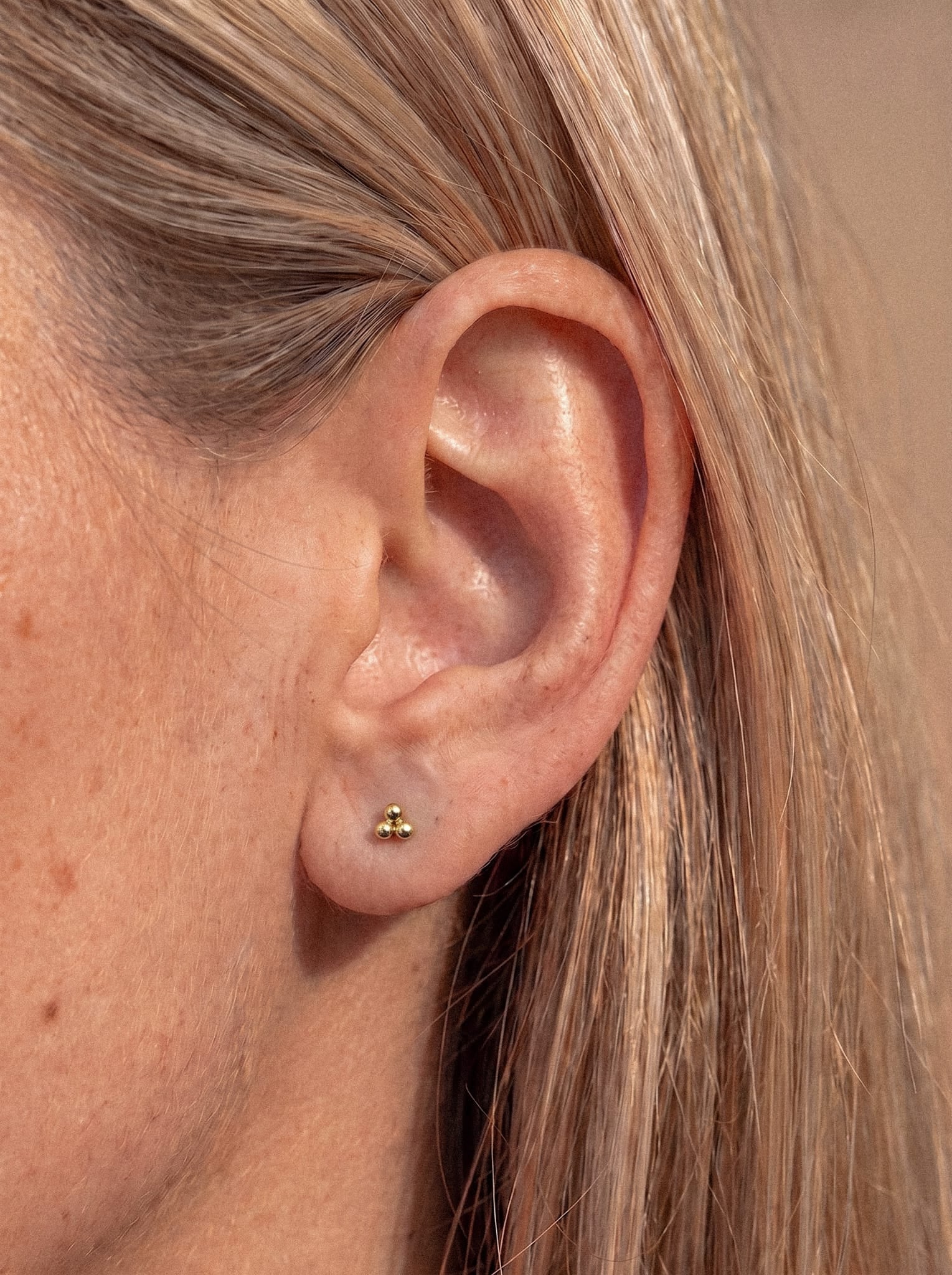 Gros plan sur l'oreille d'une personne, où l'on aperçoit des cheveux blonds raides. L'oreille est ornée, au niveau du lobe, d'une petite boucle d'oreille « Trinity Liso » en or de la marque Titanio-Noble Art, mettant en valeur une boucle d'oreille « dorado » sans perçage, au design moderne et dotée d'un système de vis interne.