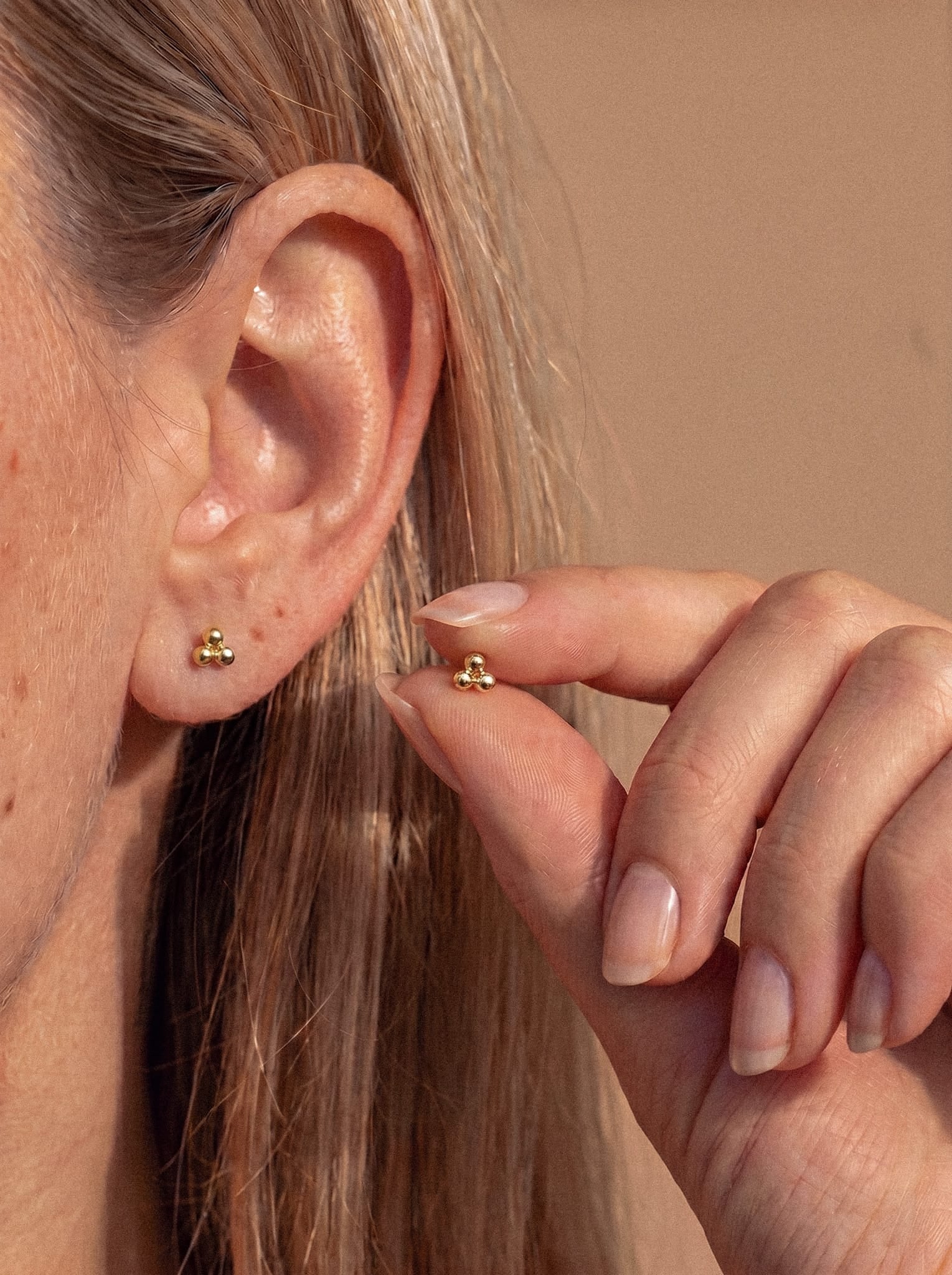 Gros plan sur l'oreille d'une personne portant la boucle d'oreille Trinity Liso de la marque Titanio-Noble Art, ornée d'un petit motif doré au design moderne. La main posée près de l'oreille tient une autre boucle d'oreille Trinity Liso dorée similaire ; les deux modèles sont dotés d'un système de vis sans fin interne garantissant une fixation sûre. La personne a les cheveux châtain clair et les ongles au naturel.