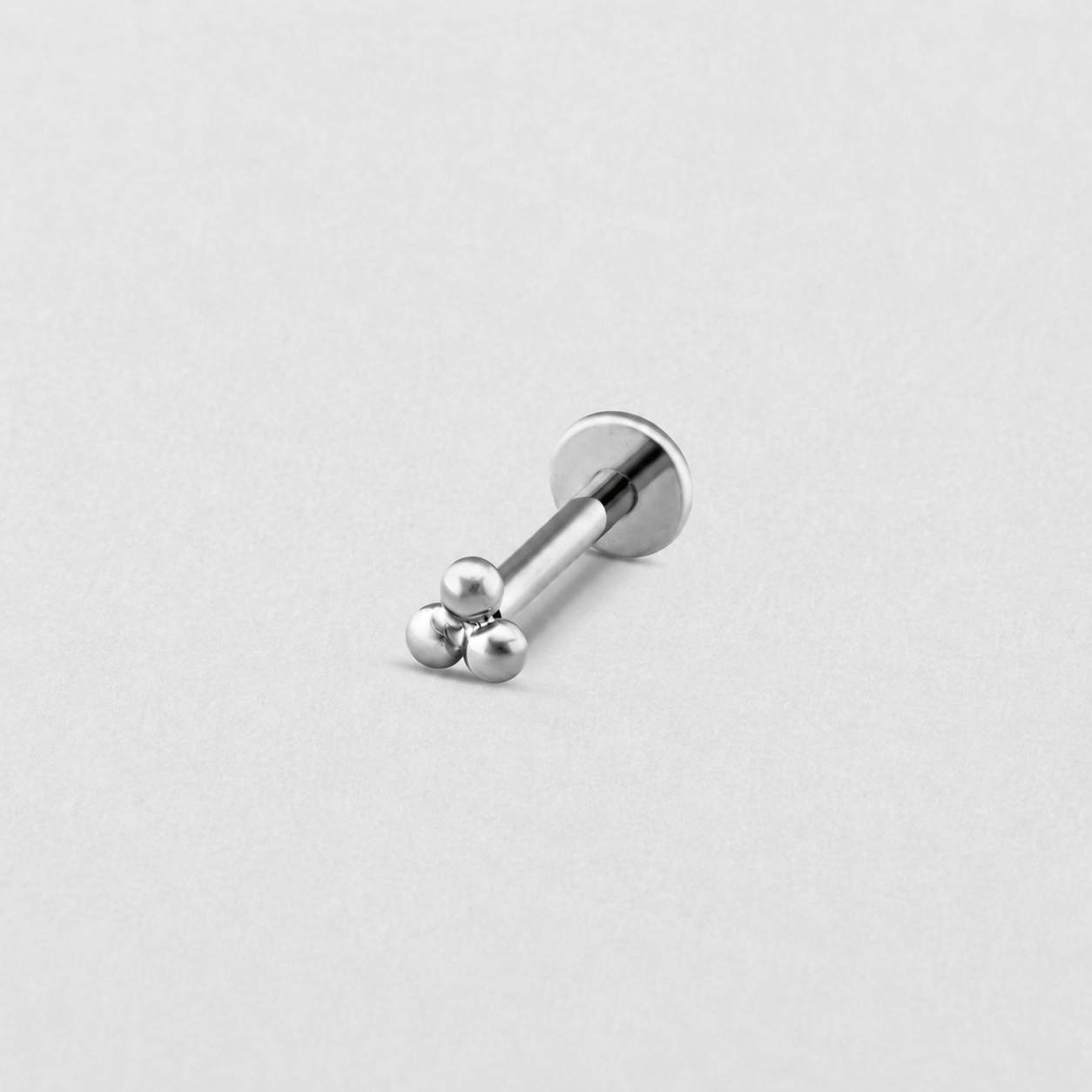 Le « Trinity Liso » de Titanio-Noble Art est un petit piercing labret en argent orné de trois minuscules boules décoratives à une extrémité et doté d'un système de filetage interne pour une fixation sûre, présenté sur un fond gris clair uni.