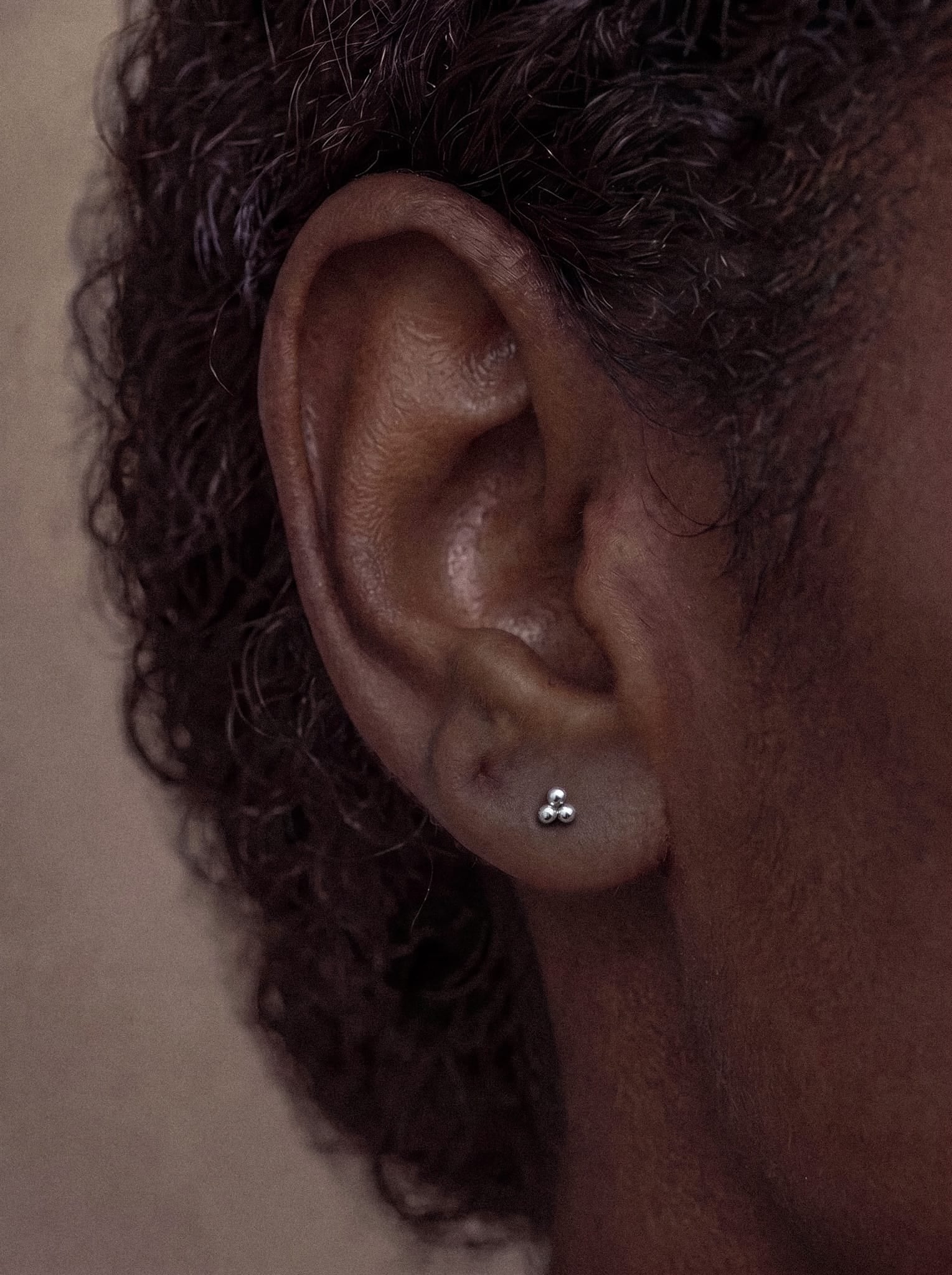 Gros plan sur l'oreille d'une personne aux cheveux bouclés, portant la boucle d'oreille « Titanio - Noble Art Trinity Liso » : un petit bijou en argent en forme de trois points reliés entre eux, doté d'un système de vis interne pour une fixation sûre et présentant un design moderne.