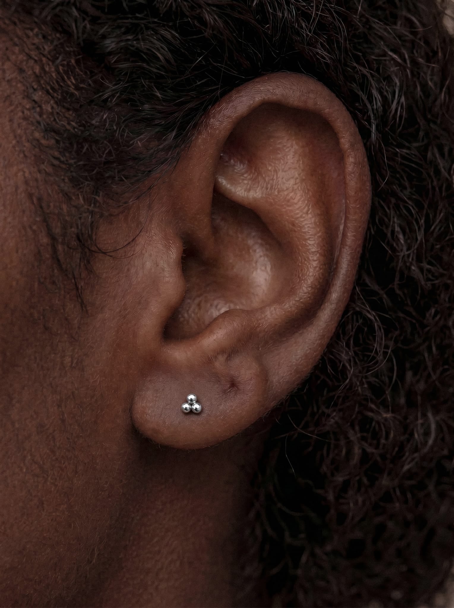 Gros plan sur l'oreille d'une personne portant la boucle d'oreille « Trinity Liso » de Titanio-Noble Art, un petit bijou en argent orné de trois points au design moderne et élégant. La peau de la personne est brun foncé, et on aperçoit des cheveux bouclés sur le côté droit de l'image. Ce piercing doré est doté d'un système de vis interne garantissant une utilisation sûre et confortable.