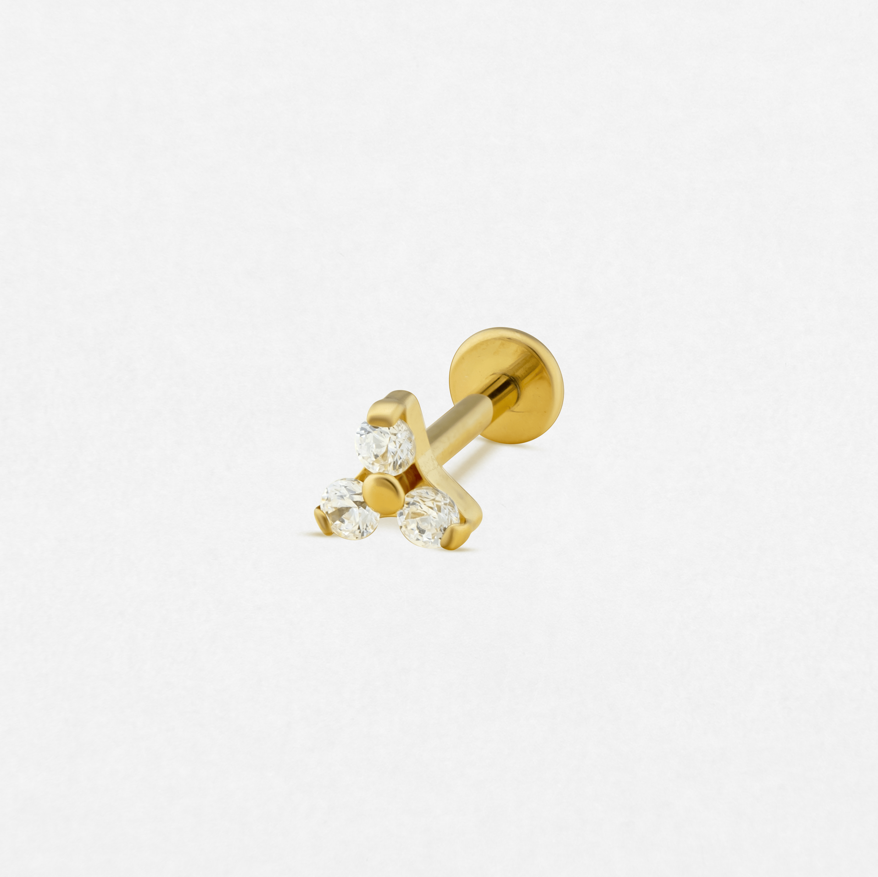 El modelo «Trinity cz blanco/oro» de Titanio-Noble Art es un pendiente de botón dorado con cierre plano y tres piedras redondas transparentes dispuestas en forma de triángulo, que combina elegancia y sencillez, y que se muestra sobre un fondo blanco liso.