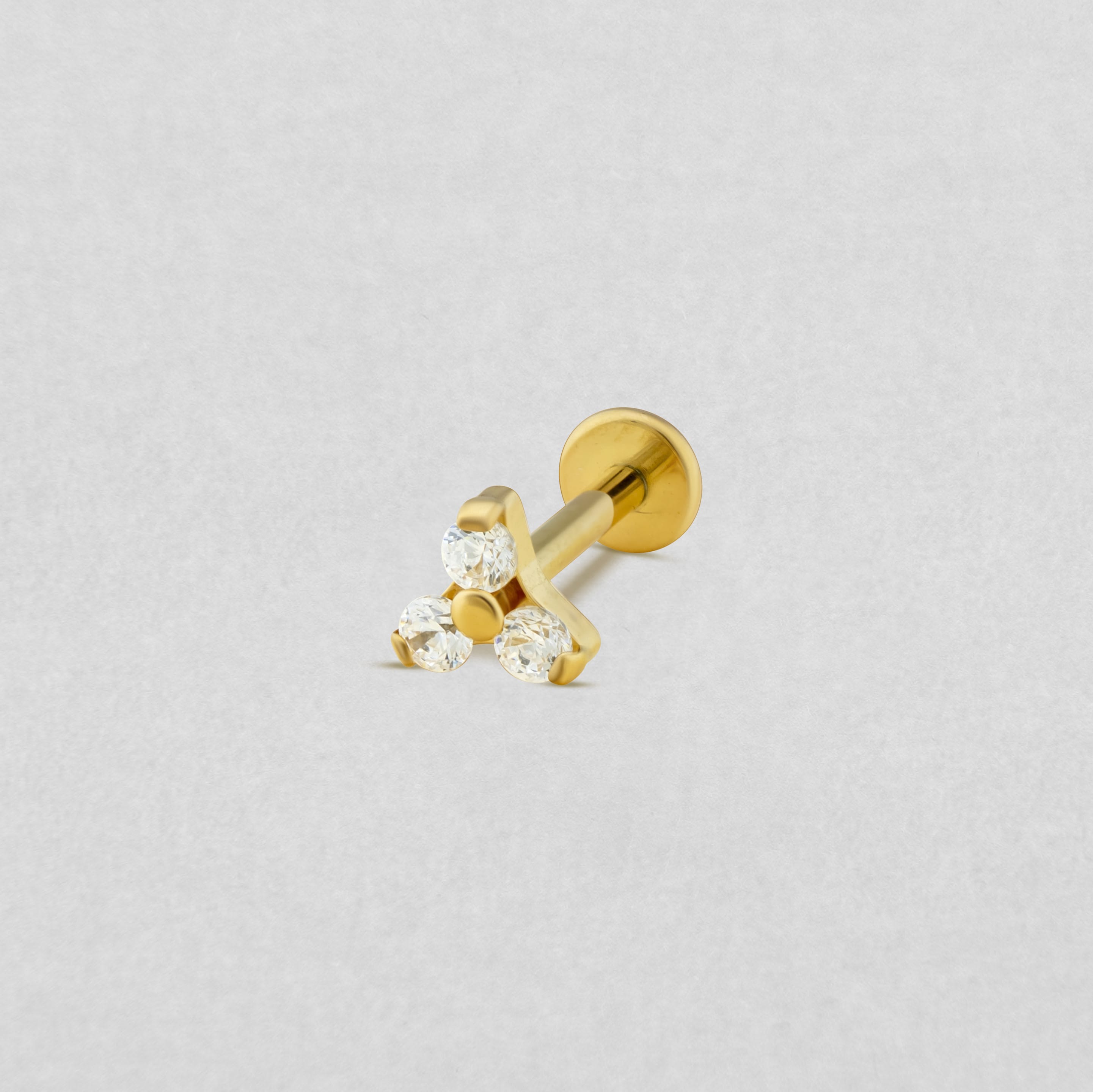 El «Titanio- Noble Art Trinity CZ» es un pendiente tipo labret de oro con tres piedras preciosas redondas transparentes dispuestas en forma de triángulo, que se muestra sobre un fondo gris claro: perfecto para tu piercing.