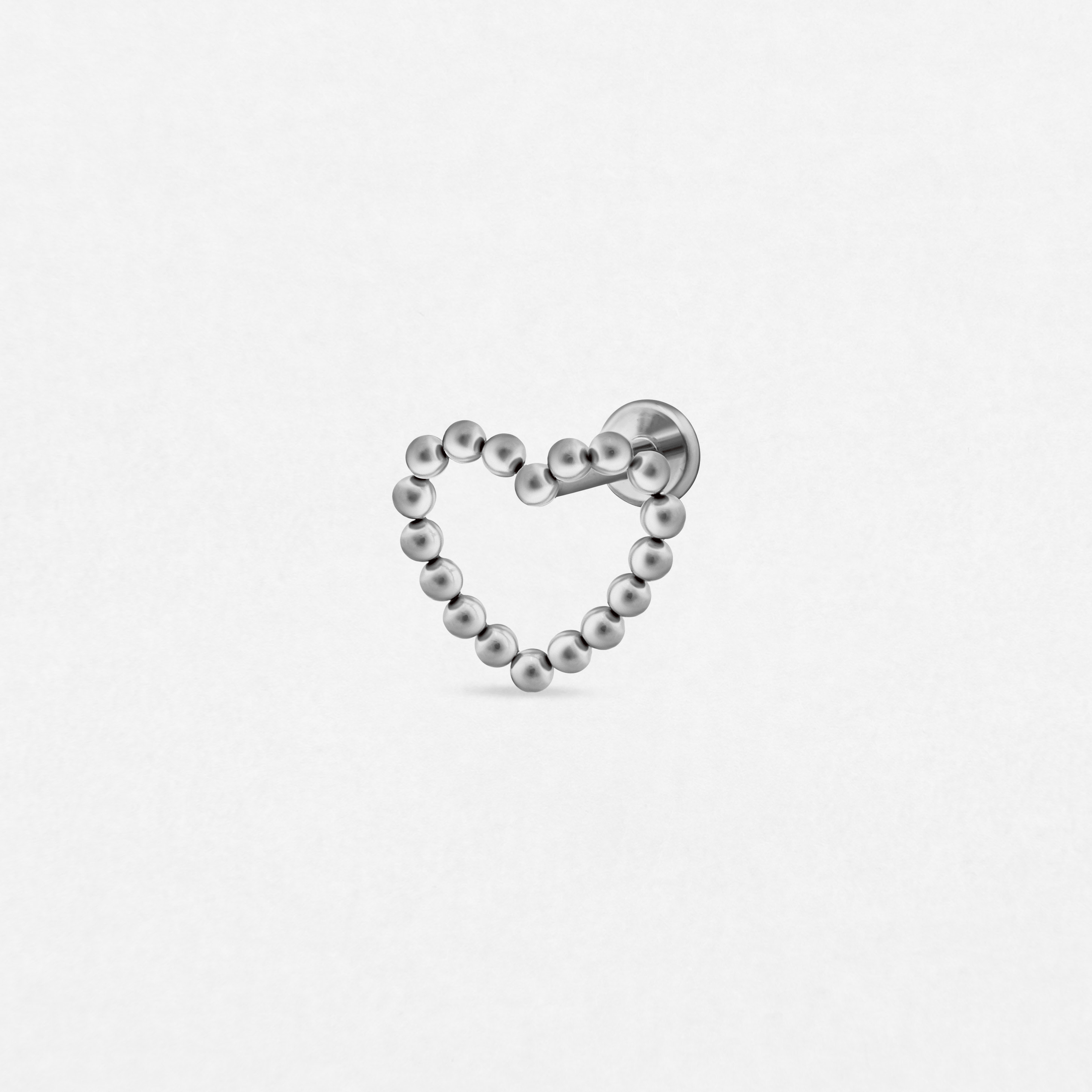 El pendiente «Amar Gold» de Titanio-Noble Art presenta una forma de corazón elaborada con diminutas cuentas metálicas dispuestas en forma de lazo y con un pasador recto. Inspirado en los piercings minimalistas, este accesorio dorado se muestra sobre un fondo blanco liso.
