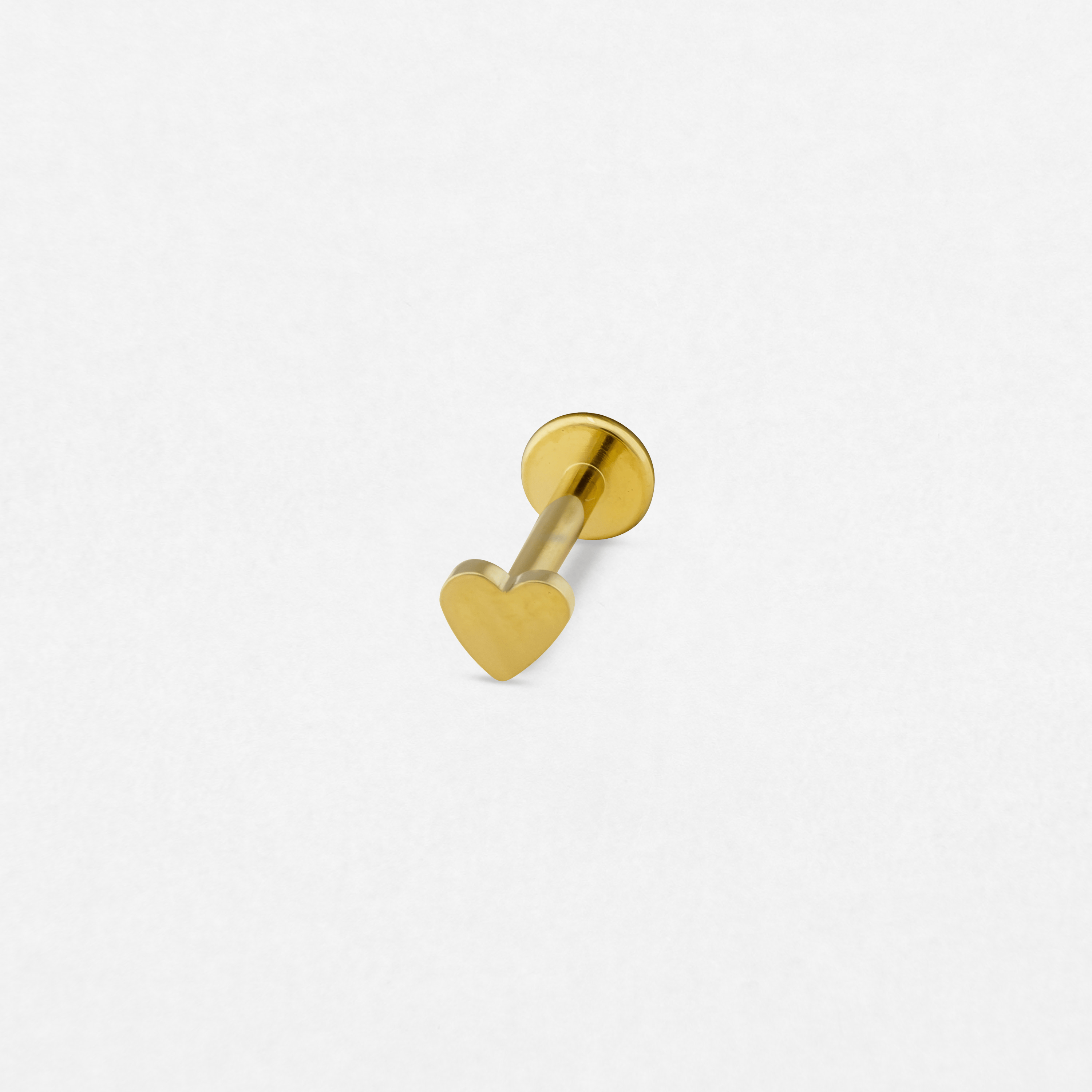 El «Love liso» de Titanio-Noble Art es un pequeño pendiente tipo barra de color dorado, con un cierre circular plano y una parte delantera en forma de corazón, que destaca sobre el fondo blanco: un refinado homenaje a la expresión personal.