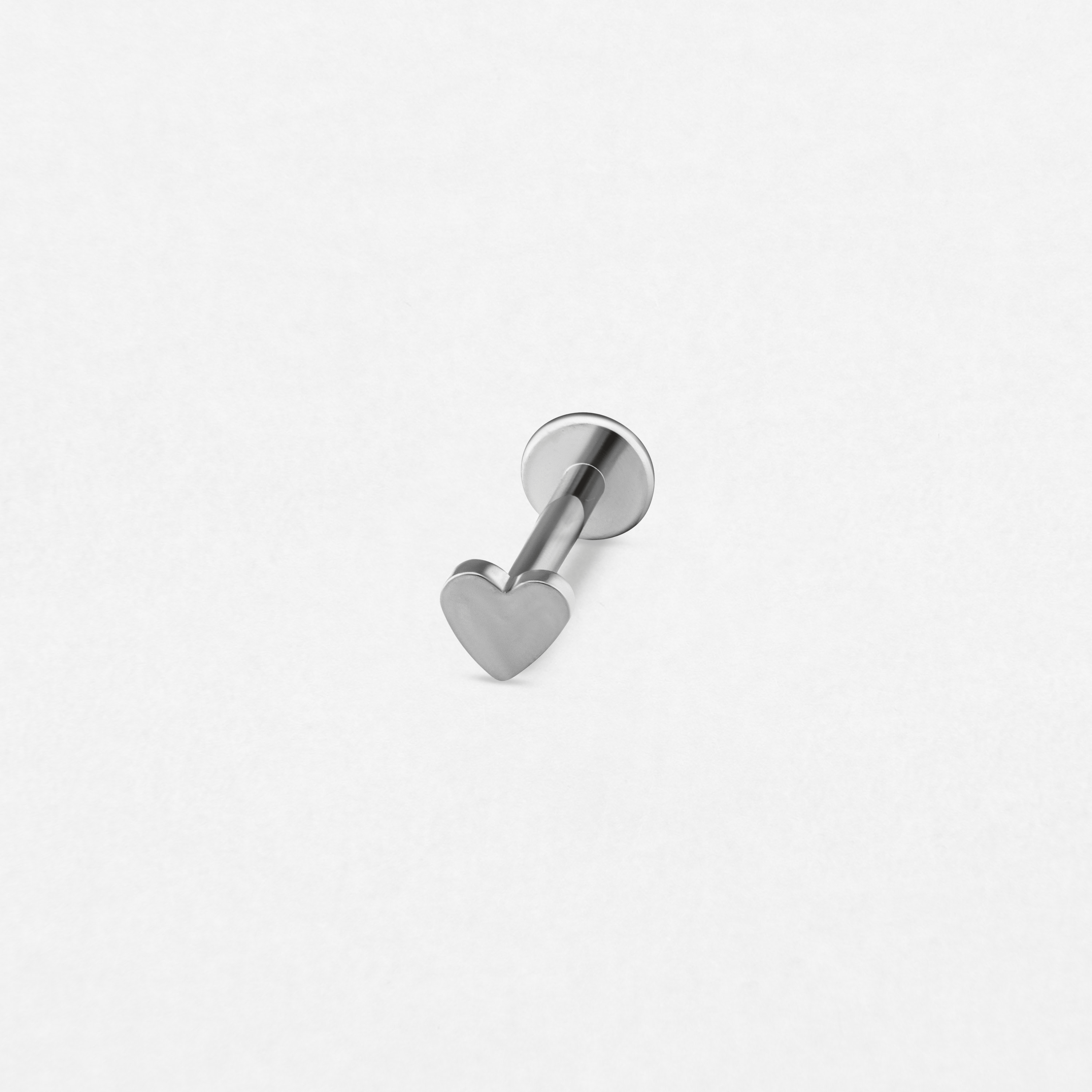 El modelo LOVE LISO SILVER de Titanio-Noble Art es un pequeño pendiente de botón de plata con un extremo plano en forma de corazón y un cierre redondo, perfecto para perforaciones recientes, que se muestra sobre una superficie blanca lisa.