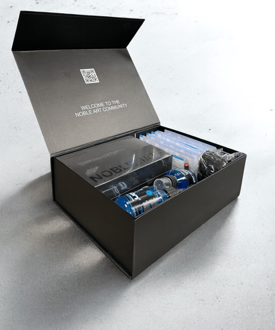Una caja negra abierta de Noble Art, con la etiqueta «Kit de tatuaje para principiantes», descansa sobre una superficie clara junto a artículos de la marca, como latas y un cuaderno negro. En el interior de la tapa, junto a un código QR, se lee: «Bienvenido a la comunidad Noble Art».