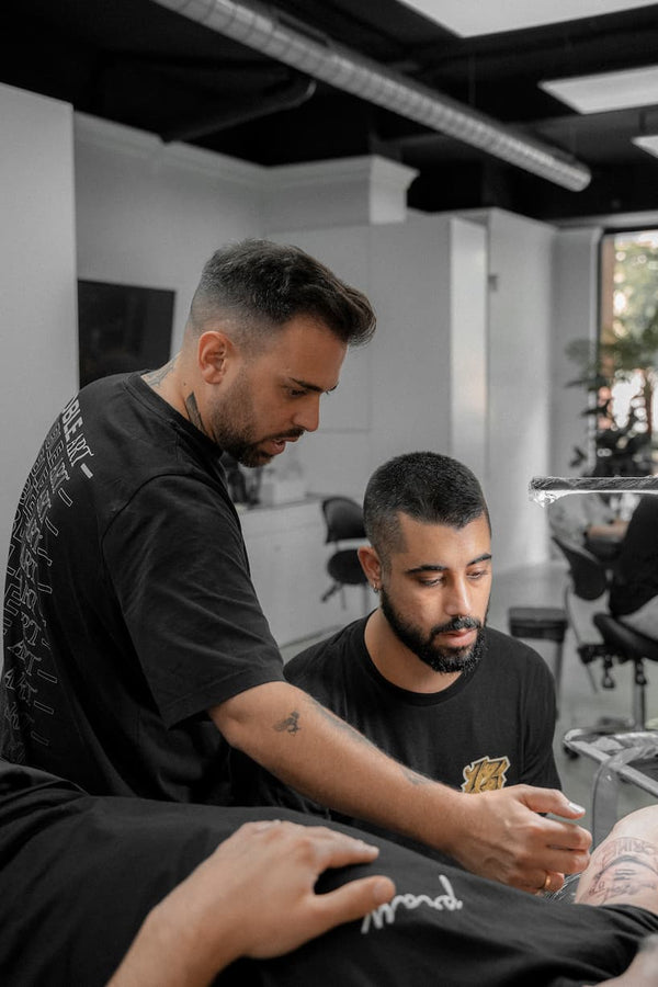 Two men in a modern tattoo studio: one is using a tattoo machine on a client's arm while the other assists. The professional setting suggests expertise in tattooing and possibly piercing or laser removal services.