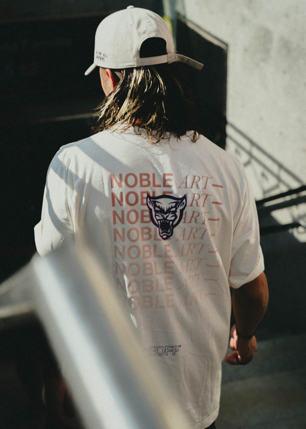 Eine Person mit langen Haaren trägt eine weiße Kappe und ein weißes T-Shirt mit der Aufschrift «NOBLE ART» und einer Wolfszeichnung auf dem Rücken.