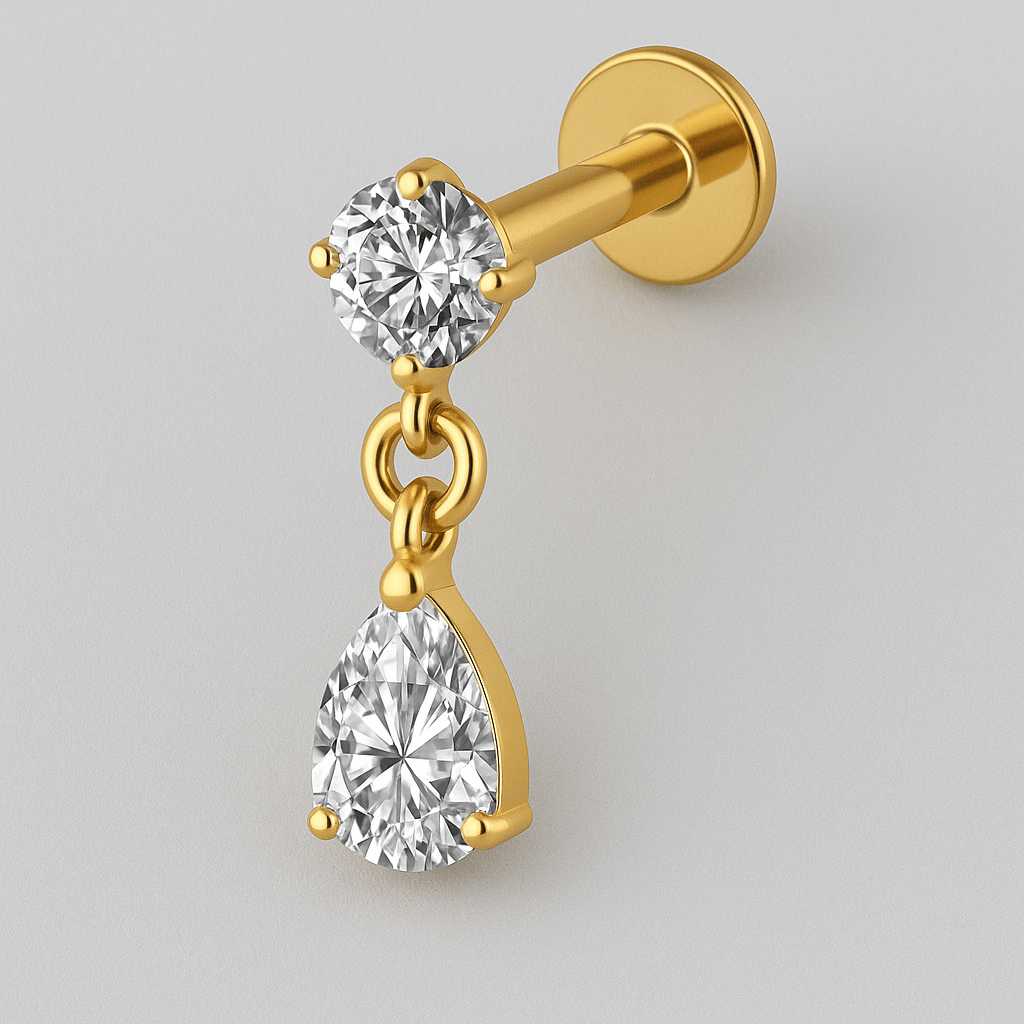 El pendiente de oro LIA de NOBLE ART BEAUTY SOCIEDAD LIMITADA cuenta con un pendiente de diamante redondo y un diamante colgante en forma de pera, ambos engastados en oro, perfectos para cualquier tipo de piercing y fotografiados sobre un fondo gris claro.