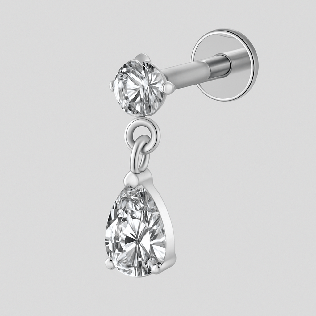 El pendiente de botón LIA de NOBLE ART BEAUTY SOCIEDAD LIMITADA presenta un diamante de talla redonda con un diamante colgante en forma de pera, ambos engastados con garrafas minimalistas, sobre un fondo liso de color gris claro: una opción elegante para cualquier tipo de piercing.