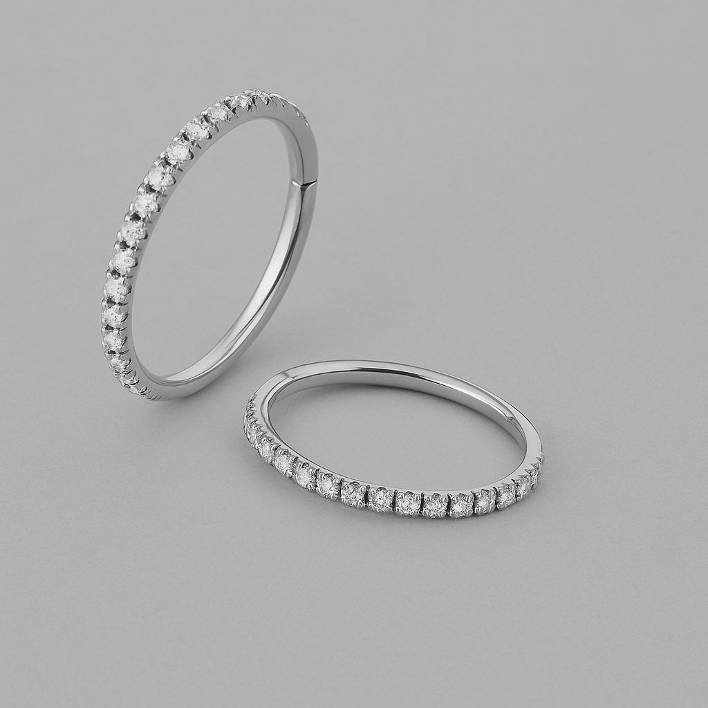Los anillos EMILY WHITE de NOBLE ART BEAUTY SOCIEDAD LIMITADA presentan finas bandas de plata con pequeños diamantes blancos redondos a lo largo de la mitad superior, que se muestran sobre un fondo gris claro; son perfectos para combinarlos en un estilo minimalista o para lucirlos junto con tu piercing favorito.