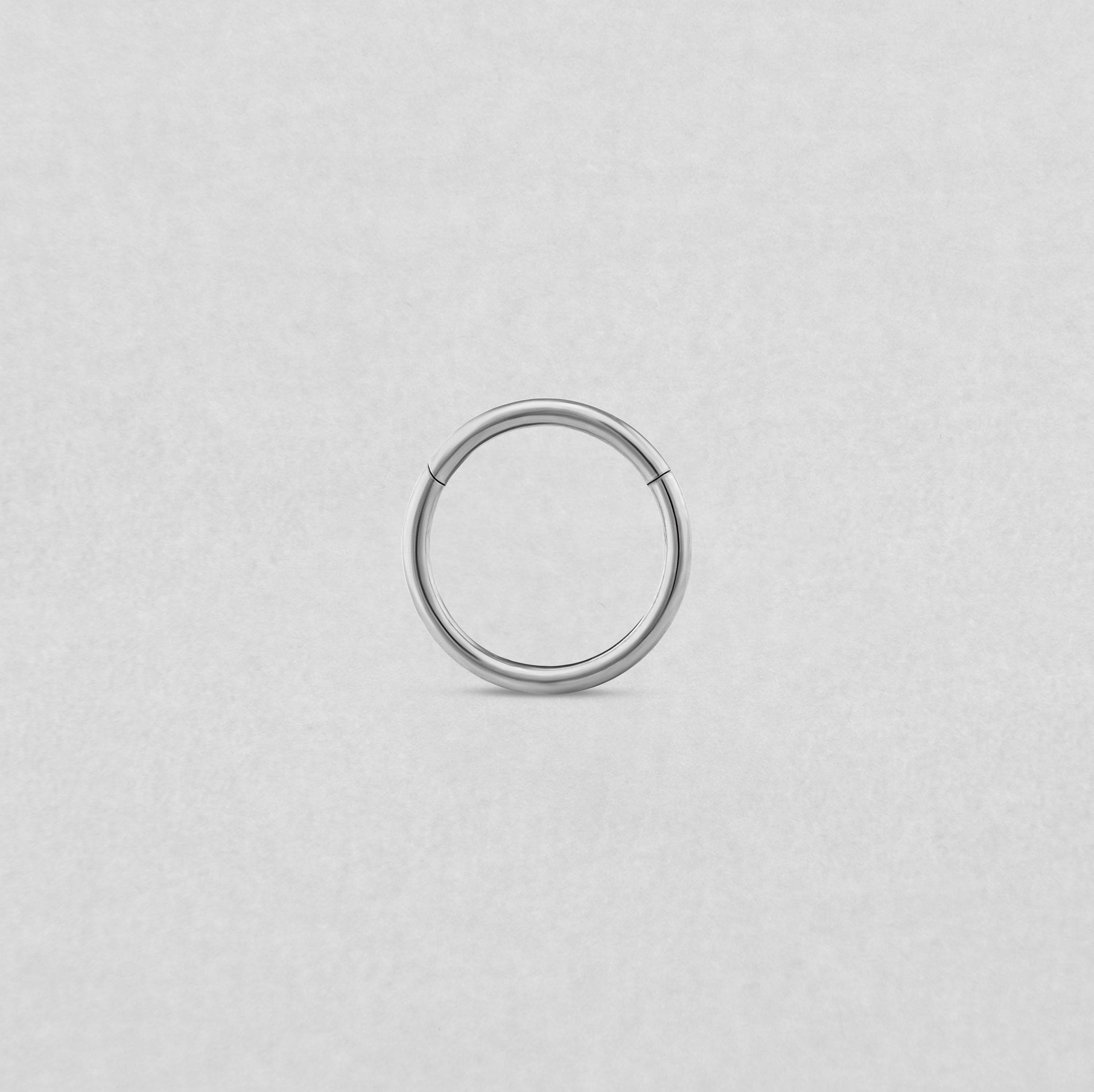 Un sencillo pendiente de aro «Clicker Basico» de plata de NOBLE ART BEAUTY SOCIEDAD LIMITADA aparece en el centro de un fondo liso de color gris claro, con una pequeña abertura en la parte superior que sugiere que se trata de un aro con bisagra o sin costuras.
