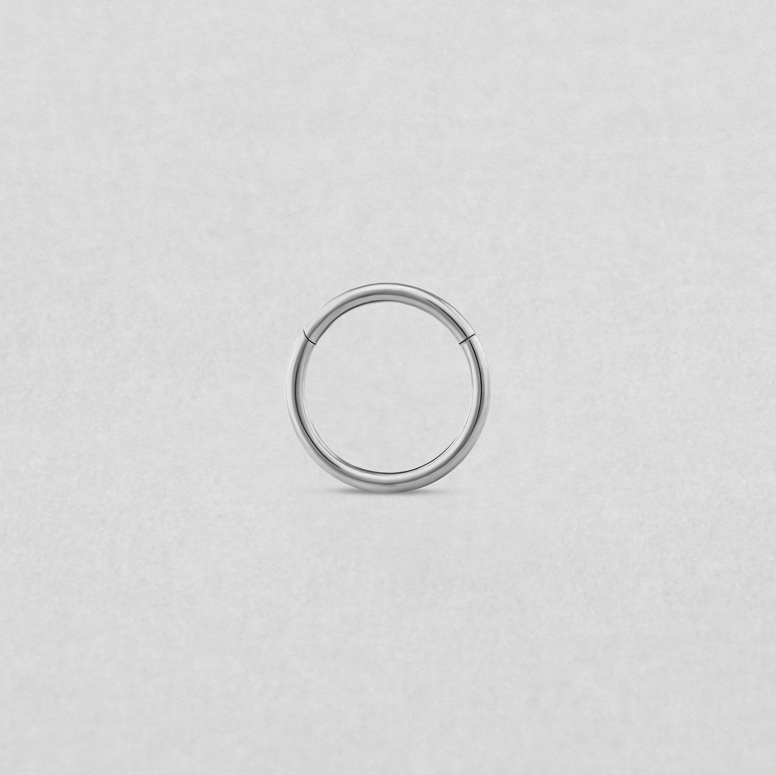 Un sencillo pendiente de aro «Clicker Basico» de plata de NOBLE ART BEAUTY SOCIEDAD LIMITADA aparece en el centro de un fondo liso de color gris claro, con una pequeña abertura en la parte superior que sugiere que se trata de un aro con bisagra o sin costuras.